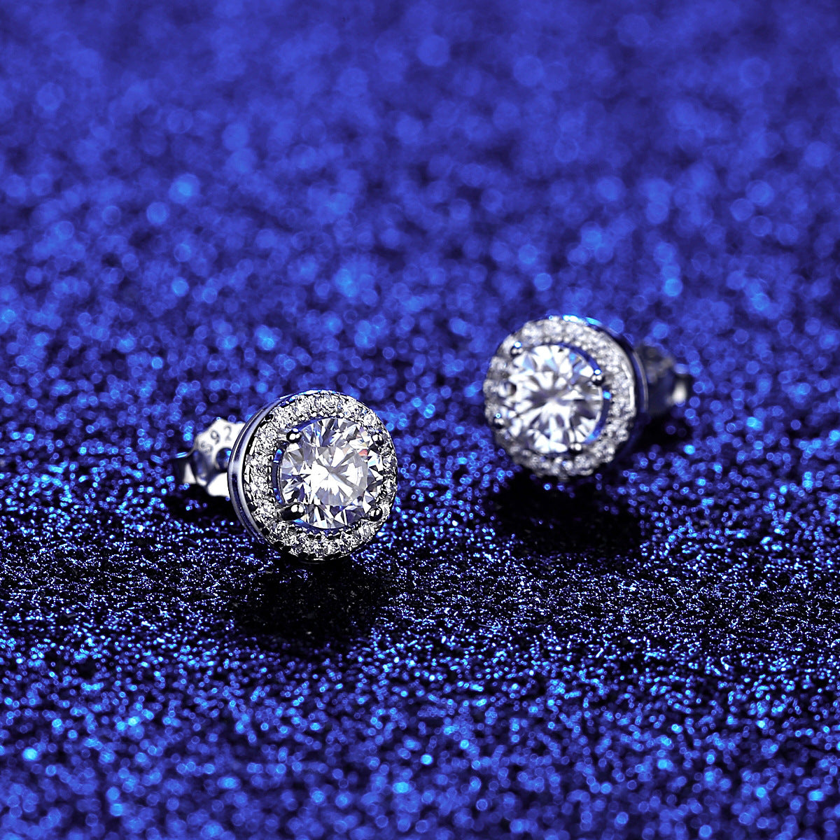 A pair of sparkling silver earrings with round, clear gemstones on a blue glitter background.
